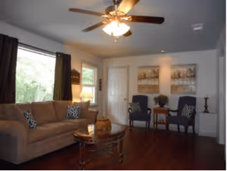 A cozy living room with a beige sofa adorned with patterned pillows, a wooden coffee table with decorative items, two blue armchairs with patterned cushions, a small wooden side table with a plant, two framed artworks on the wall, a ceiling fan with lights, and windows with dark curtains letting in natural light.