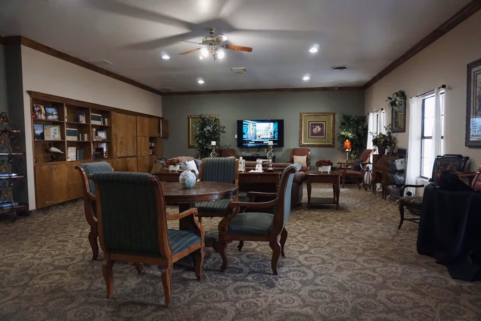 Community lounge with upholstered chairs and tables facing a wall-mounted TV and bookshelves.