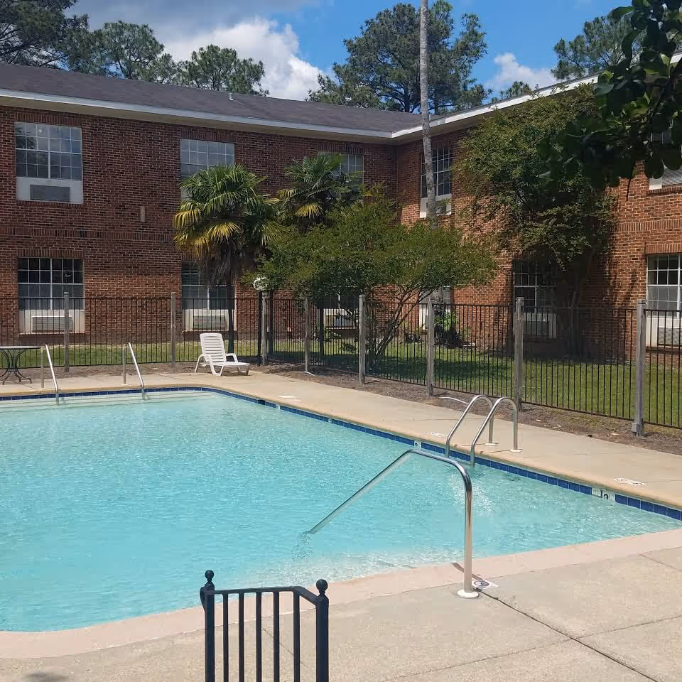 Outdoor swimming pool with clear blue water in front of a two-story red-brick building and fenced courtyard.