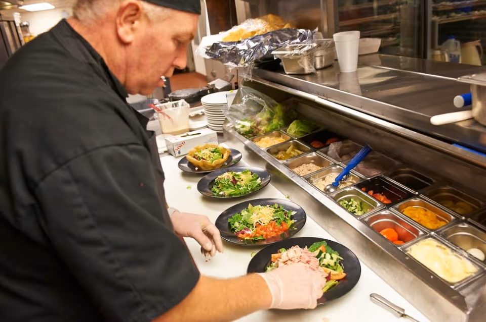A chef wearing a black uniform and gloves is preparing several plates of fresh salads in a kitchen with various salad ingredients in metal containers on the counter.