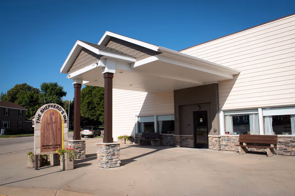 Exterior view of Shepherd's Inn assisted living facility with a covered entrance supported by two columns, stone and siding facade, benches near the entrance, and a wooden sign displaying the facility's name.