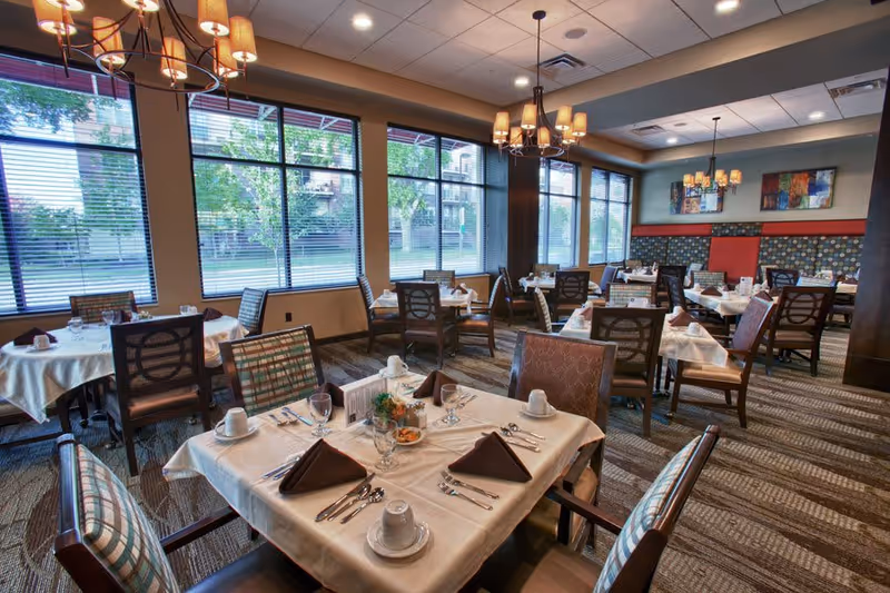 A well-lit dining room with multiple tables set for meals, featuring white tablecloths, folded brown napkins, cups, glasses, and silverware. The room has large windows with blinds, patterned carpet, and decorative chandeliers hanging from the ceiling. Artwork and cushioned seating line the walls.