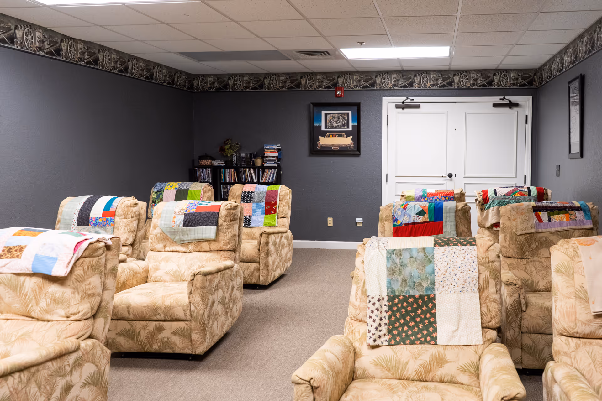 A cozy room with several beige patterned armchairs arranged in rows, each with a colorful patchwork quilt draped over the back. The walls are painted gray with a decorative border near the ceiling. A bookshelf filled with books and DVDs is in the corner, and framed artwork hangs on the walls. Double white doors are at the back of the room.