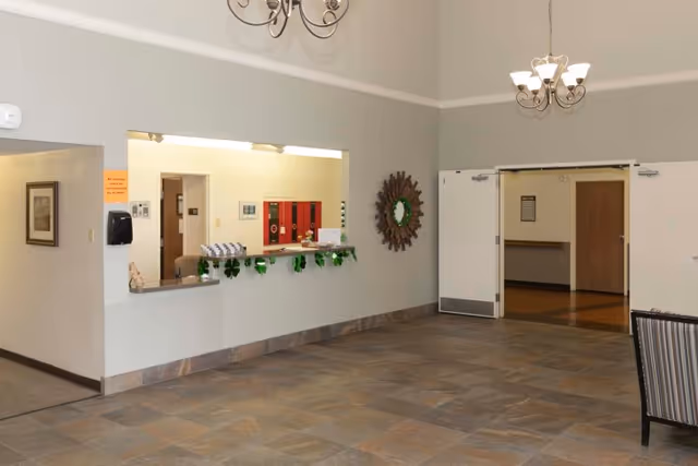 Interior view of a reception area in a senior living facility with a counter window decorated with green garlands, a wall mirror with a sunburst frame, double doors leading to another room, and a chandelier hanging from the ceiling.