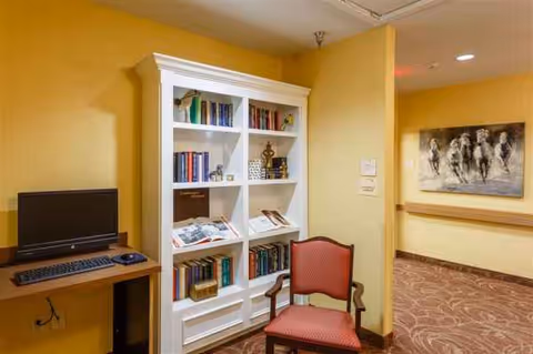 A white bookshelf filled with books stands beside a desk with a computer in yellow-walled room.