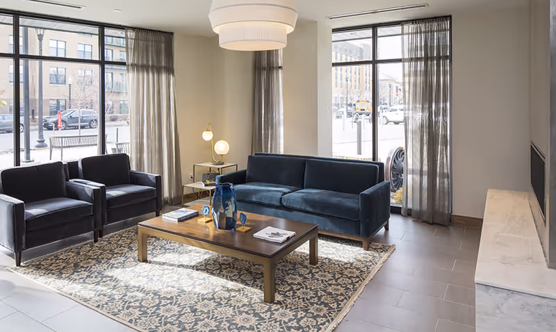 Bright modern lounge with a blue sofa, two armchairs, a wooden coffee table on a patterned rug and large windows.