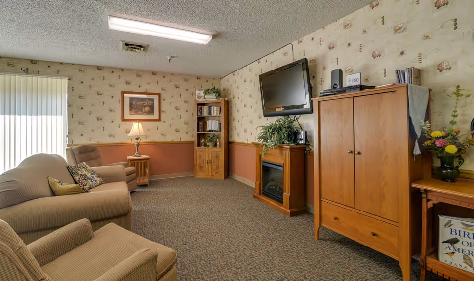 Cozy common living room with upholstered chairs and sofa, a wall-mounted TV over a fireplace, wooden cabinets and bookshelf, and a side table with a lamp.