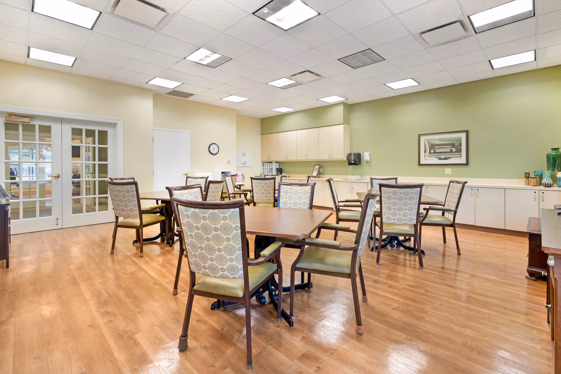 Bright dining/activity room with multiple tables and upholstered chairs, wood floors, cabinets along a green wall, and glass double doors.