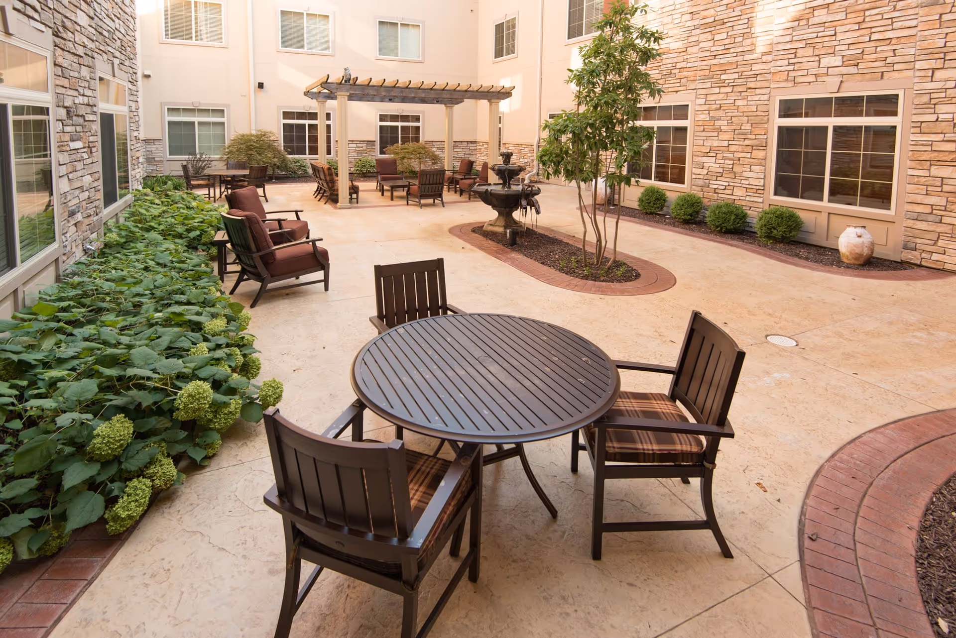 Outdoor courtyard area with a round table and four chairs in the foreground, cushioned chairs arranged under a wooden pergola, a small fountain, and various plants and shrubs along the edges. The courtyard is surrounded by a building with stone and beige walls and multiple windows.