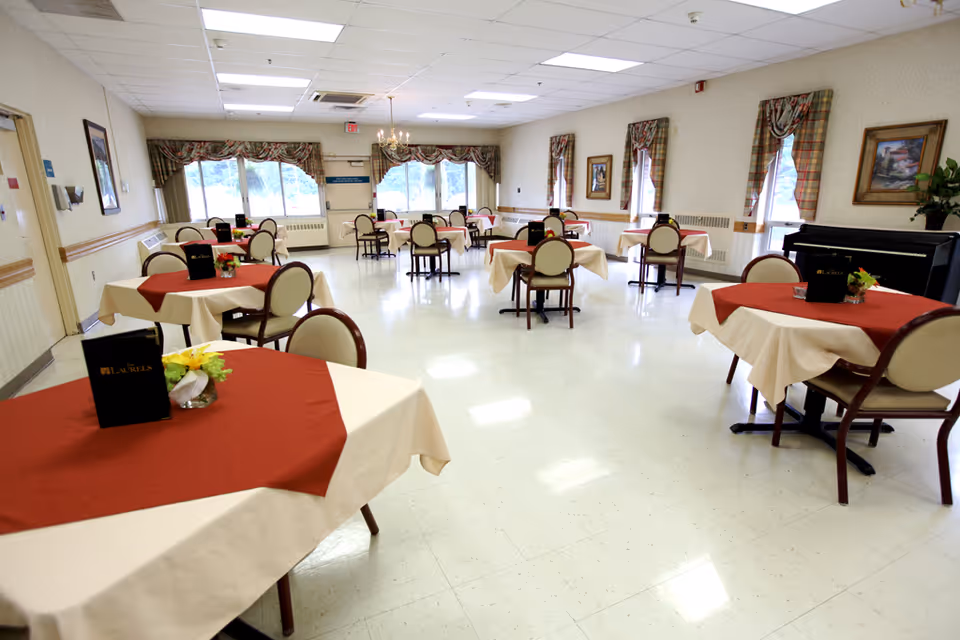 A spacious dining room with multiple round tables covered with beige tablecloths and red table runners. Each table has a small flower arrangement and a menu labeled 'Laurels'. The room has large windows with plaid curtains, framed paintings on the walls, and a piano in the corner. The floor is light-colored and the ceiling has fluorescent lighting.