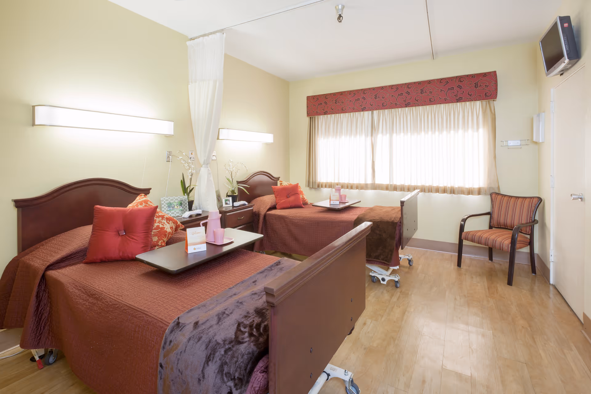 A bright and clean room with two hospital-style beds covered in brown bedspreads and red pillows. Each bed has a small overbed table with a pink cup and a card. There is a nightstand with a telephone and decorative plants between the beds. A large window with beige curtains and a red valance lets in natural light. A striped armchair is placed near the window, and a small wall-mounted television is in the upper right corner. The floor is wooden, and the walls are painted light yellow.