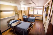 Shared hospital room with two beds, curtains, and wood flooring.