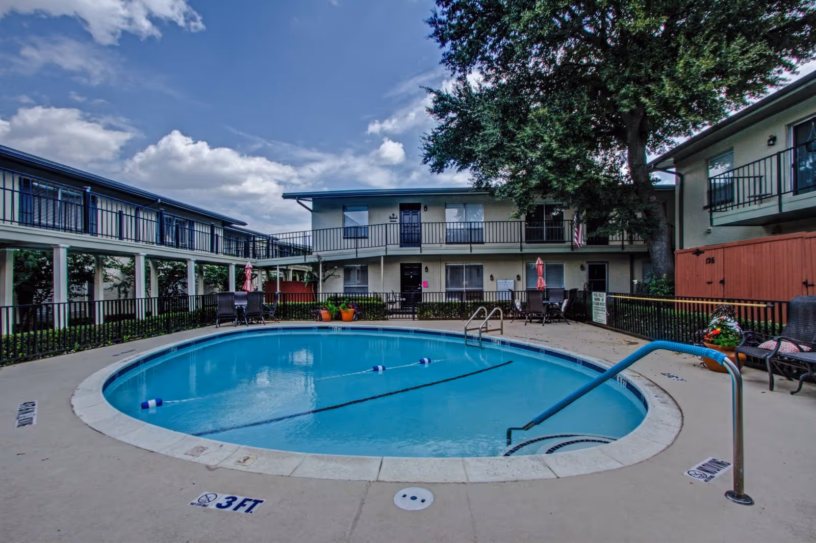 Outdoor swimming pool in the courtyard of a two-story residential building with balconies and patio furniture under a partly cloudy sky.