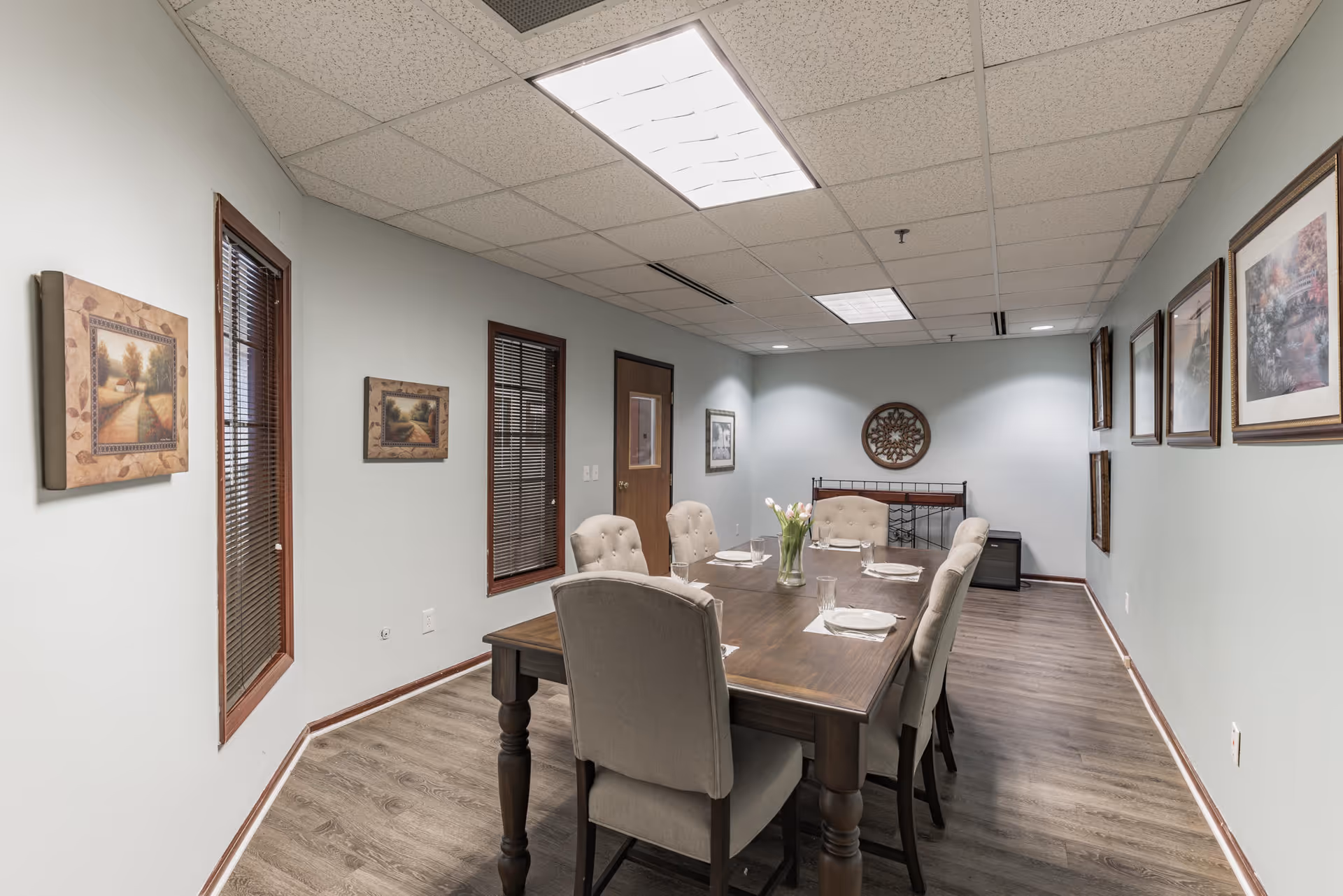 A dining room with a wooden table set for six people with plates, glasses, and a vase of flowers in the center. The room has light-colored walls adorned with framed artwork, wood-trimmed windows with blinds, and a wooden floor. The ceiling has recessed lighting panels.