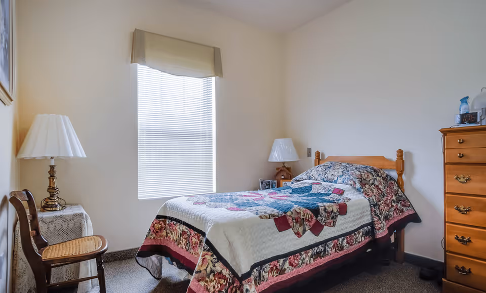 A small bedroom with a single wooden bed covered with a floral and patchwork quilt. There is a wooden dresser with drawers on the right side, a bedside table with a lamp and a clock, and a wooden chair with a woven seat on the left. A window with closed blinds and a beige valance is centered on the wall behind the bed.