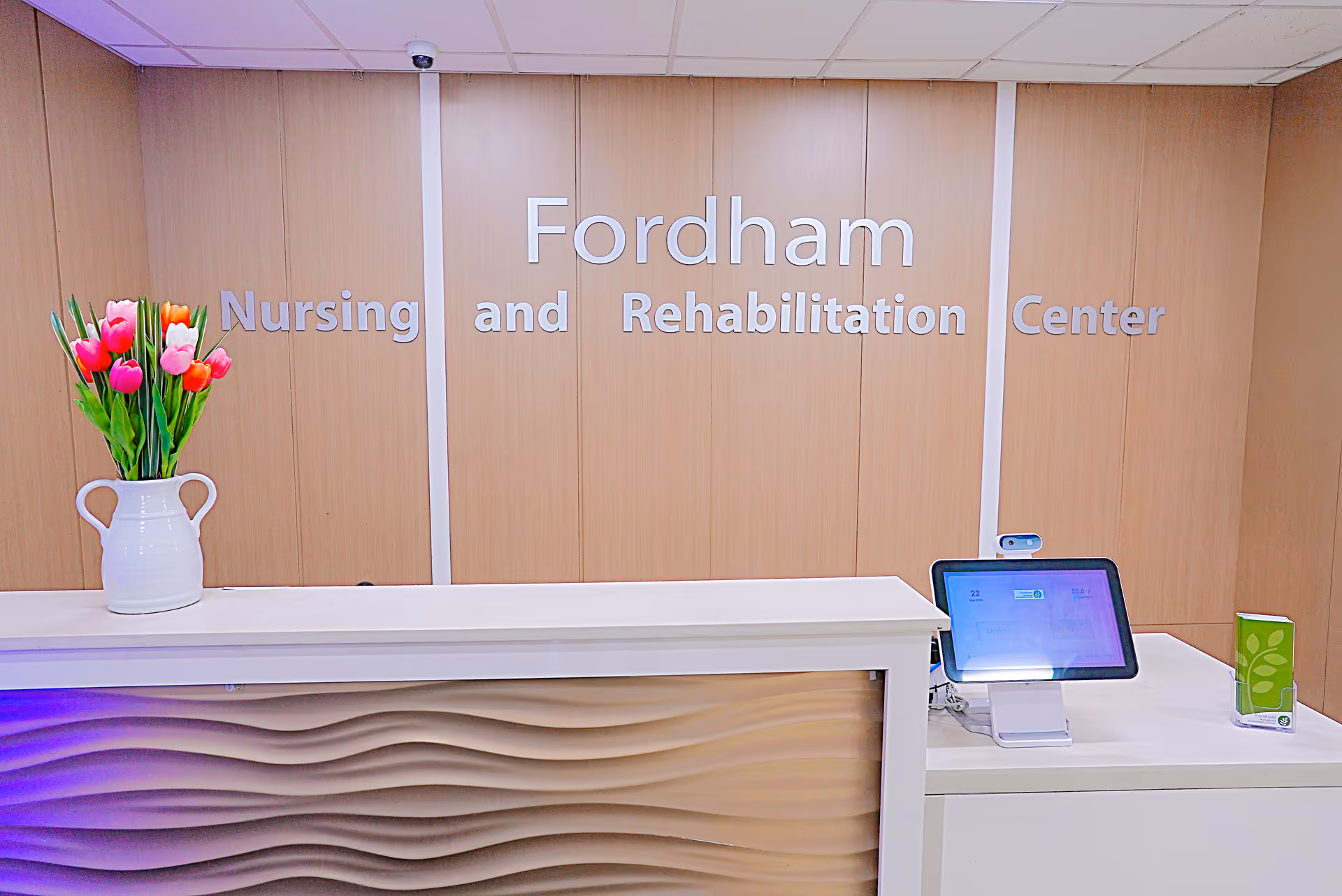 Reception desk with a vase of tulips and a tablet in front of a wall sign reading "Fordham Nursing and Rehabilitation Center".