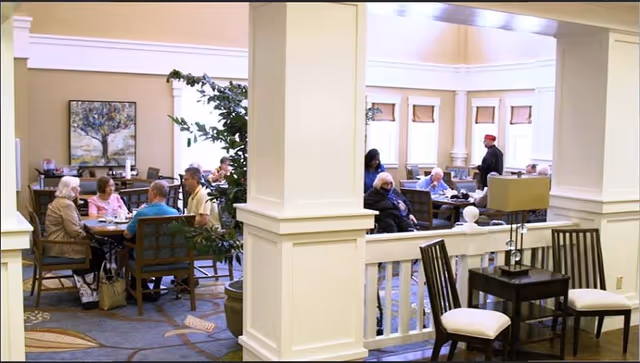A common area in a senior living facility with several elderly people sitting at tables and conversing. The room has beige walls, large windows with blinds, and decorative plants. There are wooden chairs and tables, a side table with a lamp, and a painting on the wall.
