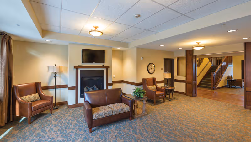 A cozy senior living facility common area with two leather armchairs and a leather loveseat arranged around a small wooden table with a plant. There is a fireplace with a mounted flat-screen TV above it, a floor lamp, a wall clock, and a staircase leading to an upper floor. The room has beige walls, a patterned carpet, and recessed lighting in the ceiling.