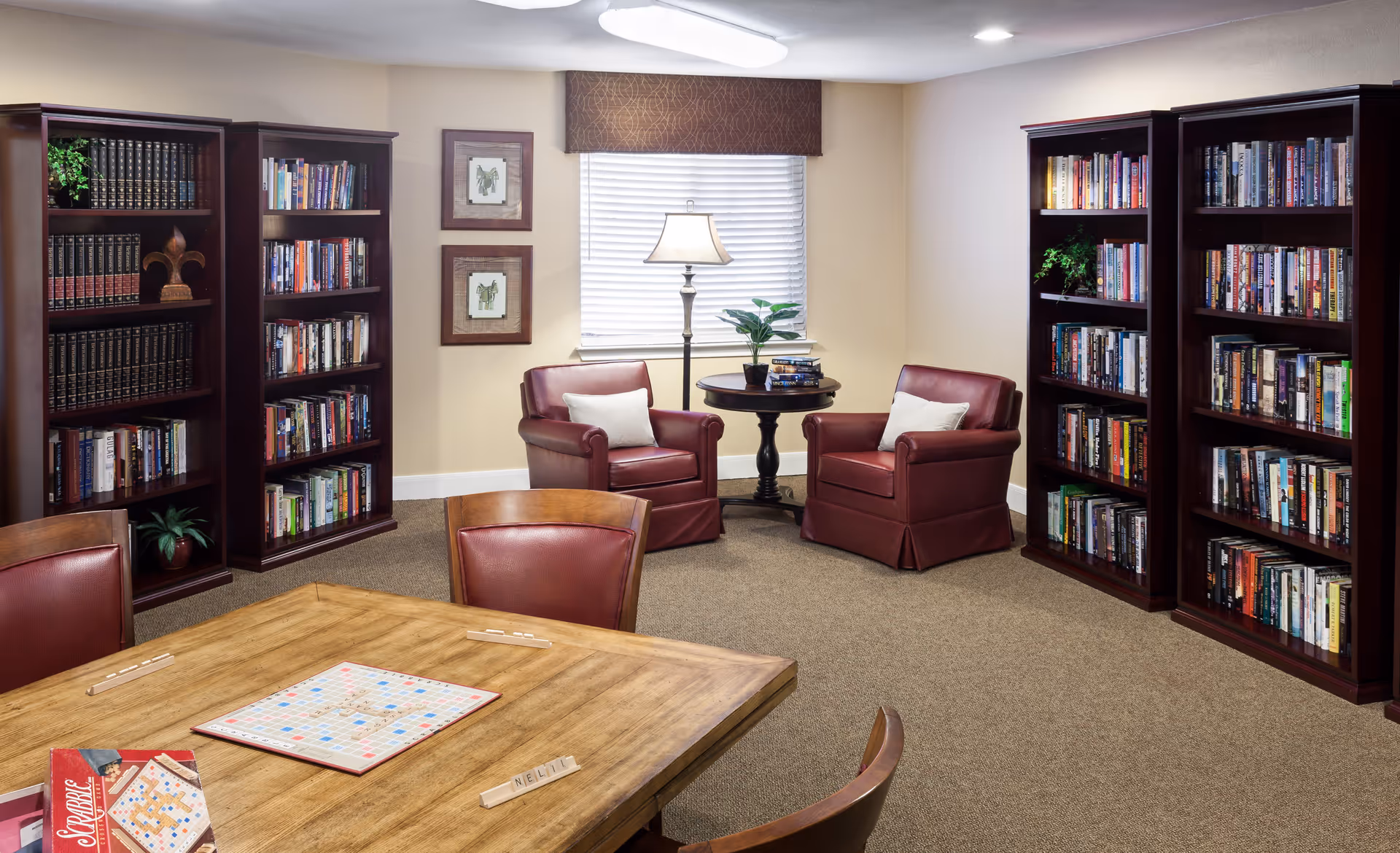 A cozy reading and game room with wooden bookshelves filled with books, two red leather armchairs with white pillows around a small round table with a lamp and plant, and a wooden table with a Scrabble board game set up in the foreground.