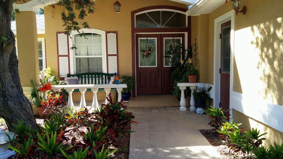 Front entrance of a yellow stucco residence featuring double red doors with wreaths, a covered porch with a bench, and landscaped plants.