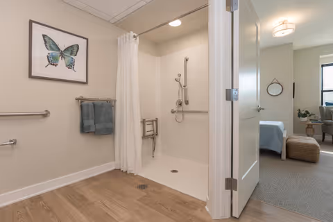 Accessible bathroom with a walk-in roll-in shower, grab bars, towel rack, and an open door leading to a bedroom.