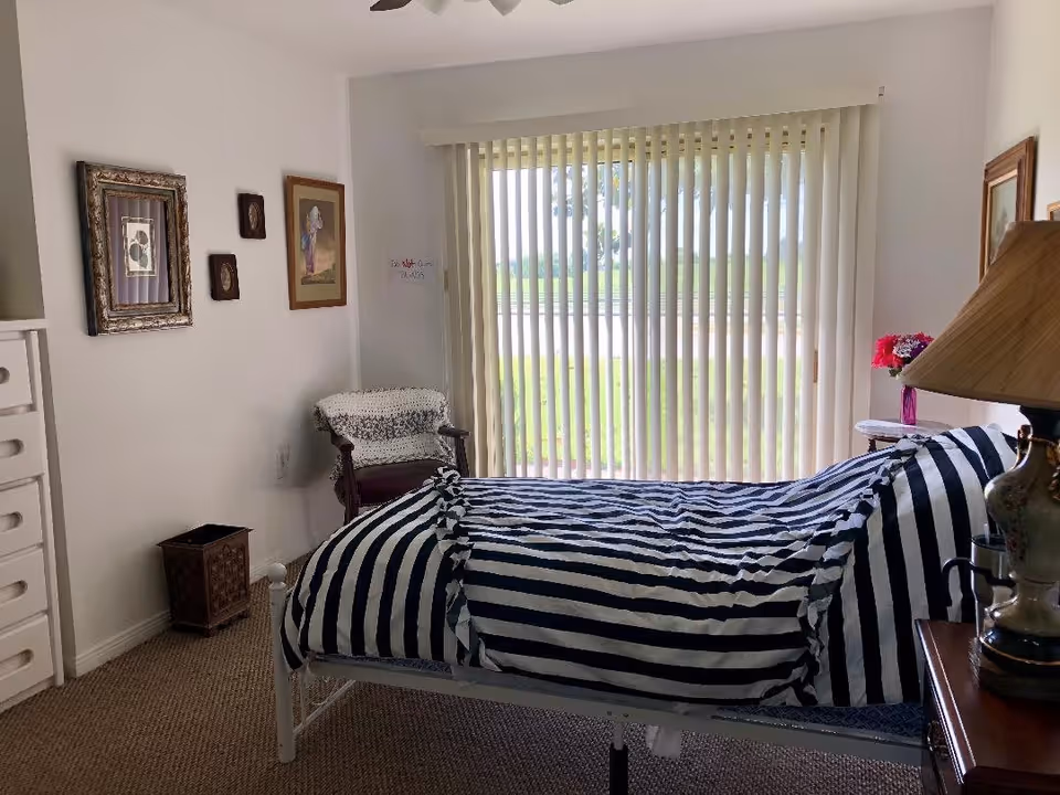 A bedroom with a bed covered in a black and white striped comforter, a wooden chair with a crocheted blanket, framed artwork on the walls, a lamp on a bedside table, and vertical blinds covering a large window overlooking a green outdoor area.