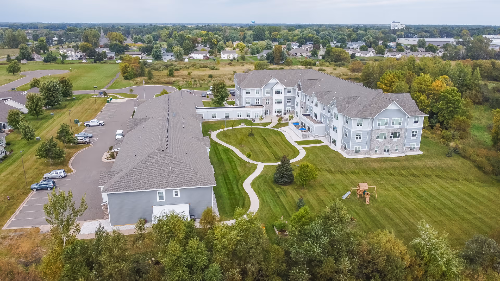 Aerial view of a senior living complex with landscaped lawns, walking paths, parking lot, and a multi-story building.