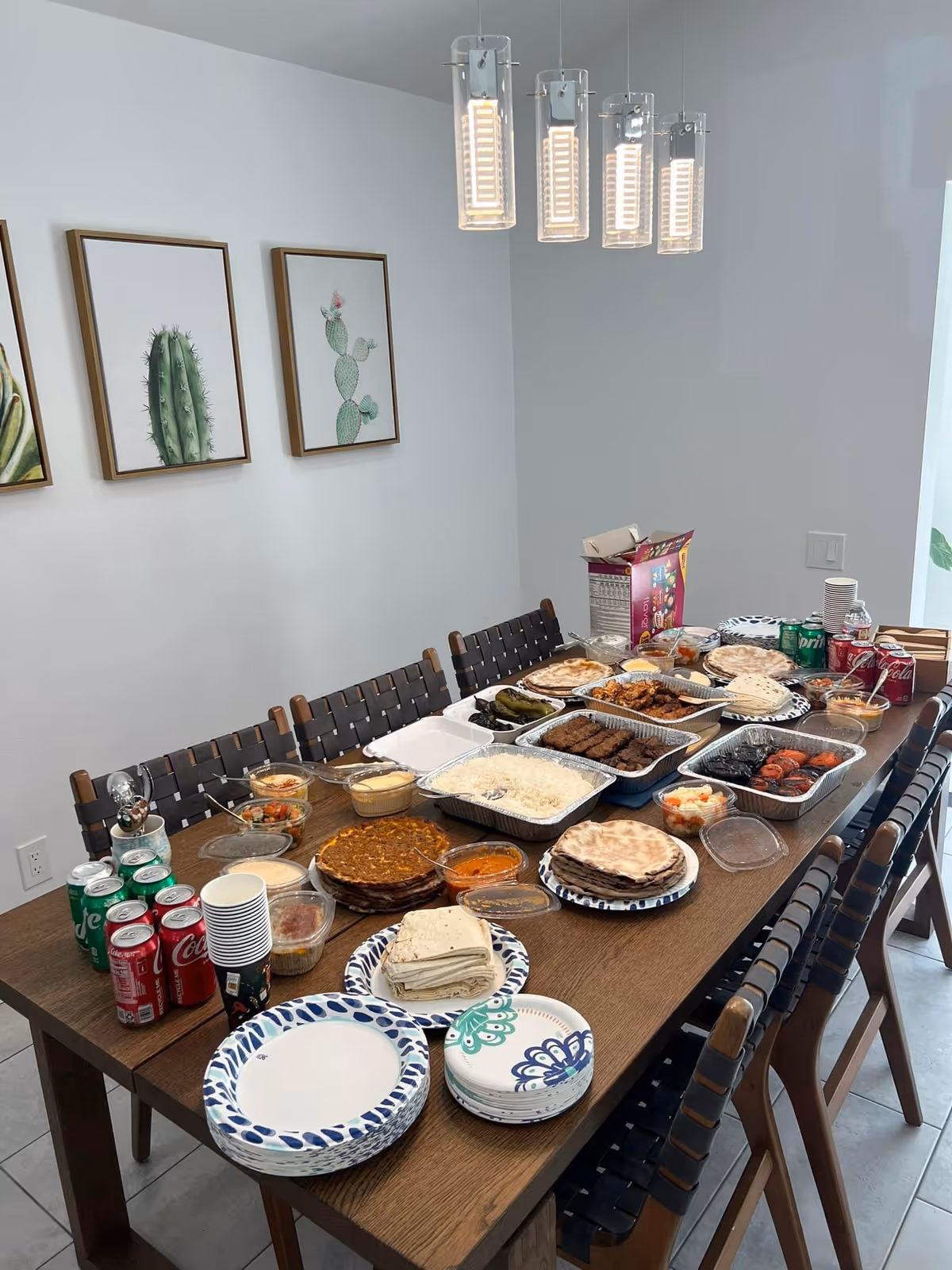 Dining room table covered with many takeout trays, plates, cans and paper cups, surrounded by chairs under pendant lights and cactus wall art.