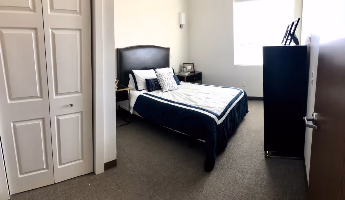 Well-lit bedroom with a navy-and-white bed, headboard, bedside tables, dresser, and closet doors.