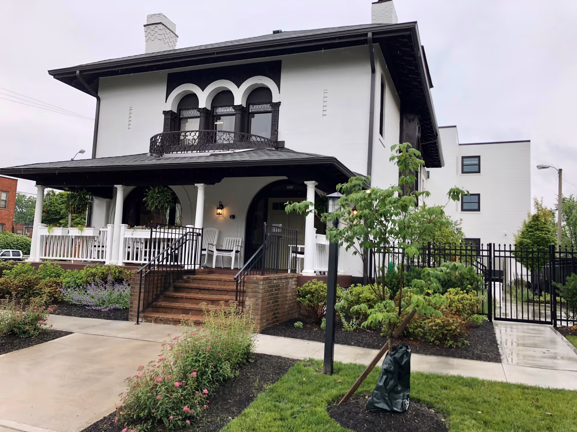Front exterior view of a two-story white building with black trim, featuring a covered porch with white columns and rocking chairs. There are steps leading up to the porch, surrounded by landscaped bushes and flowers. A small tree is planted in the foreground next to a sidewalk and a black metal fence encloses part of the property.