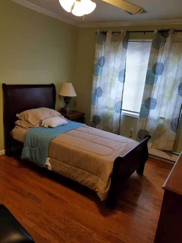 A simple single bed with wooden headboard and footboard in a bedroom with hardwood floors, a nightstand and a window with patterned curtains.