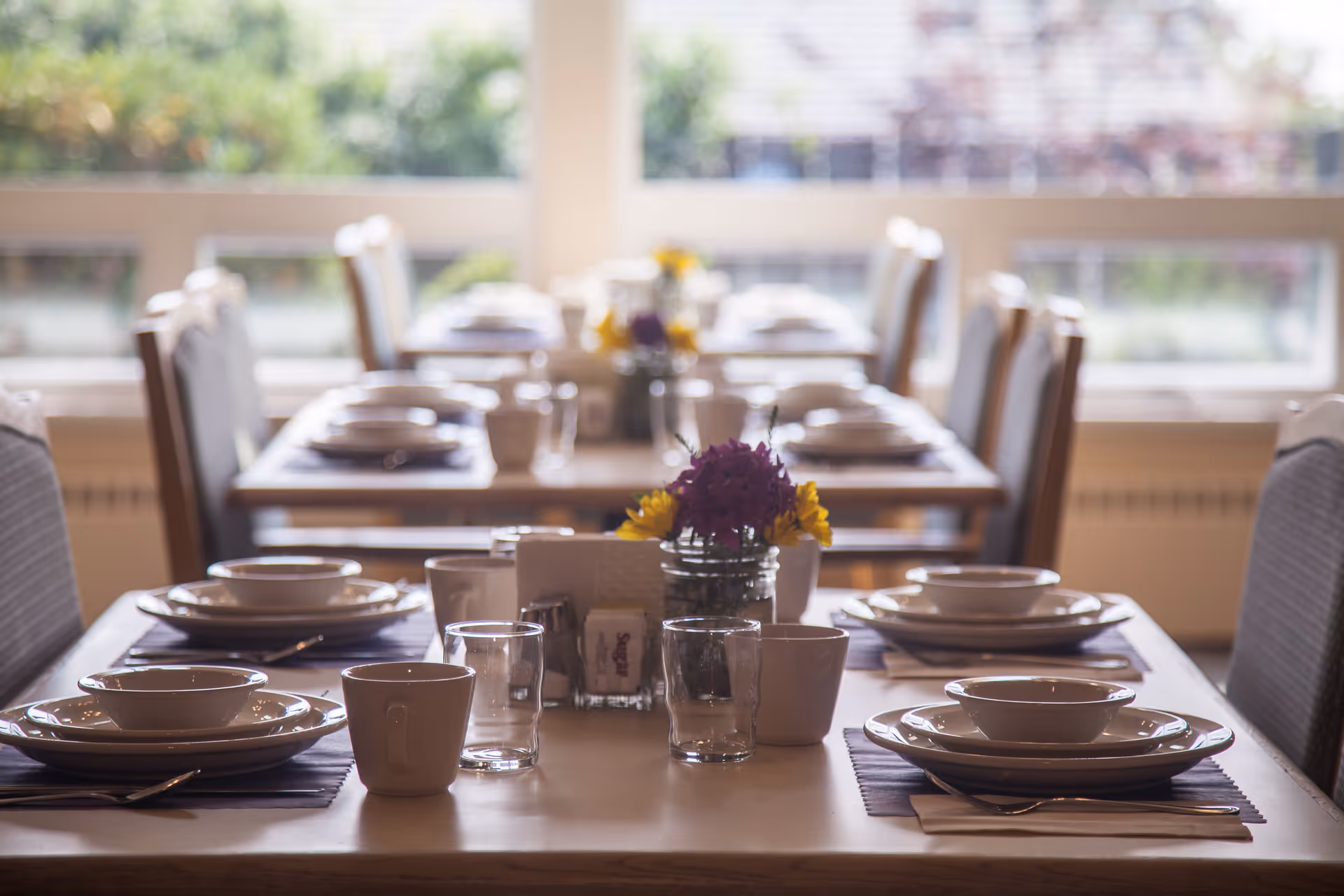 A dining table set for a meal with plates, bowls, cups, glasses, and silverware. The table has a centerpiece with a small bouquet of purple and yellow flowers. Several similar tables are visible in the background near large windows letting in natural light.