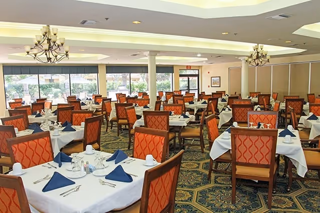 Spacious dining room with many tables set with white tablecloths, navy napkins, and patterned orange chairs under chandeliers.