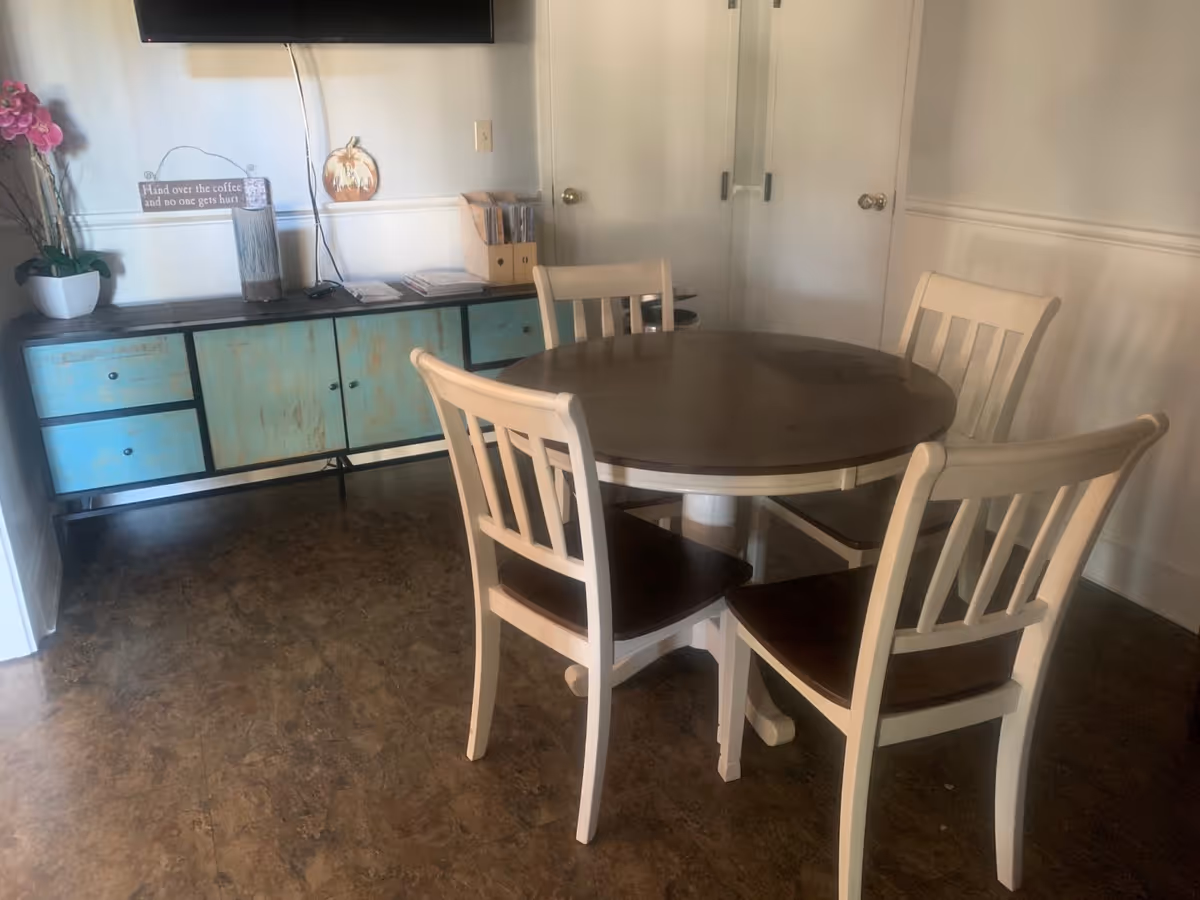 A dining area with a round wooden table and four matching chairs with white frames and dark seats. Against the wall is a rustic blue and brown sideboard with a potted orchid, a decorative sign, and some magazines or pamphlets. A flat-screen TV is mounted on the wall above the sideboard.