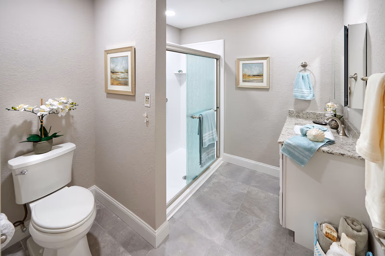 A clean and modern bathroom featuring a white toilet with a potted orchid on top, a glass-enclosed shower with a blue towel hanging on the door, a granite countertop with a sink, blue and white towels, and two framed landscape paintings on the walls.