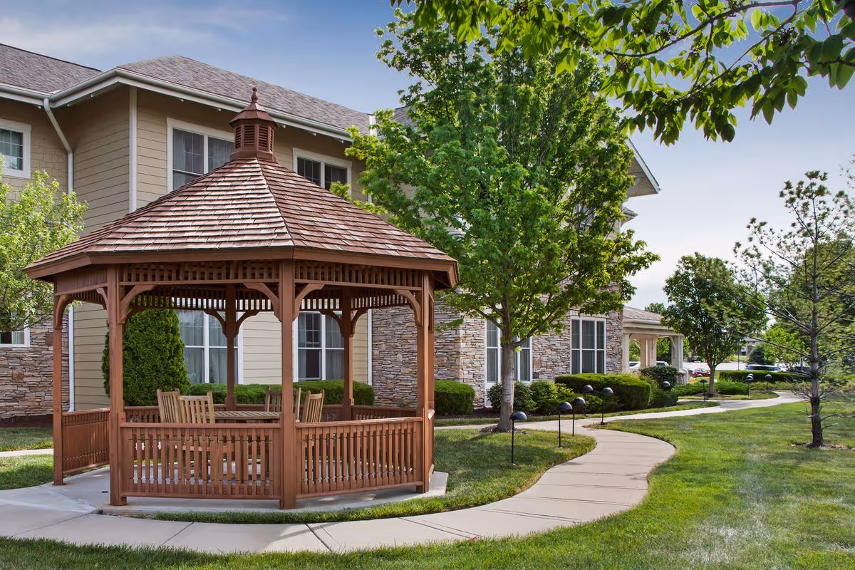 A wooden gazebo with a shingled roof and several chairs inside, situated on a concrete path surrounded by green grass and trees, with a beige and stone exterior building in the background under a clear sky.