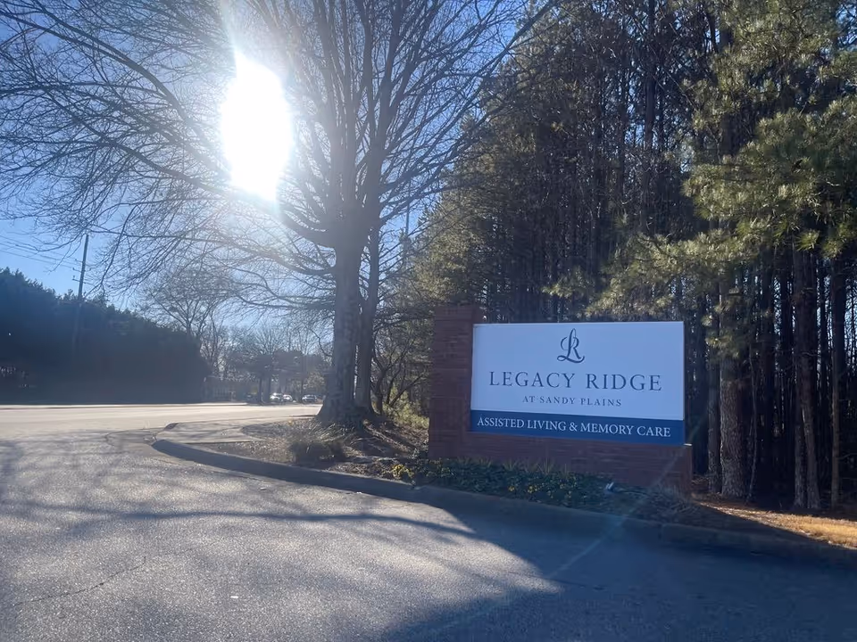 Entrance sign reading 'Legacy Ridge at Sandy Plains Assisted Living & Memory Care' by a tree-lined driveway under bright sunlight.