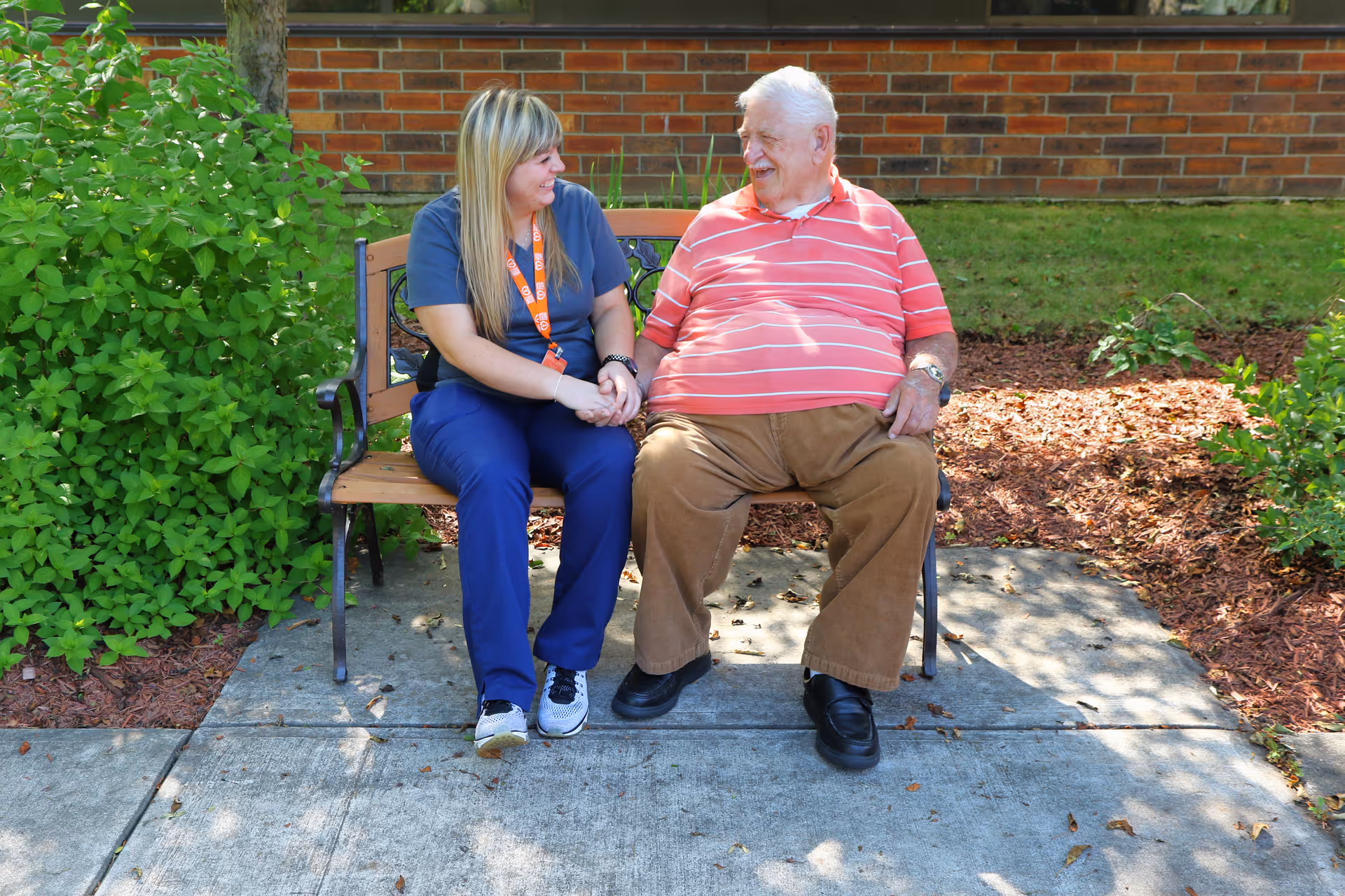 A woman in blue scrubs sitting on a wooden bench outdoors next to an elderly man wearing a red striped shirt and brown pants. They are holding hands and smiling at each other. There are green bushes and a brick wall in the background.
