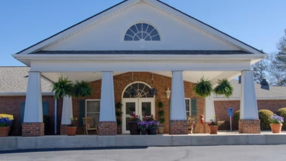 Front entrance of a brick building with a covered portico, white columns, hanging ferns, and potted flowers.