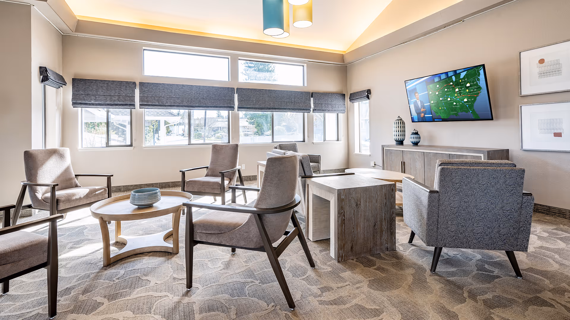 A bright and modern living room with several armchairs arranged around a round wooden coffee table and a rectangular wooden side table. Large windows with gray blinds allow natural light to fill the room. A flat-screen TV is mounted on the wall above a wooden cabinet, and two decorative vases are placed on the cabinet. The carpet has a subtle pattern, and the ceiling features recessed lighting with a soft glow.