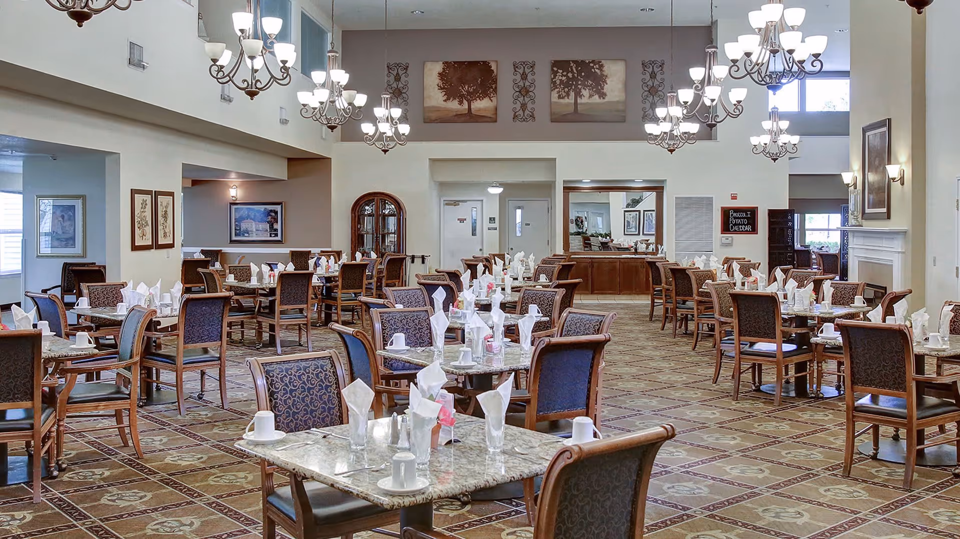 A spacious dining room in a senior living facility with multiple tables set with white napkins, cups, and glasses. The room features patterned carpet, elegant chandeliers hanging from a high ceiling, framed artwork on the walls, and a fireplace on the right side. The overall ambiance is warm and inviting.