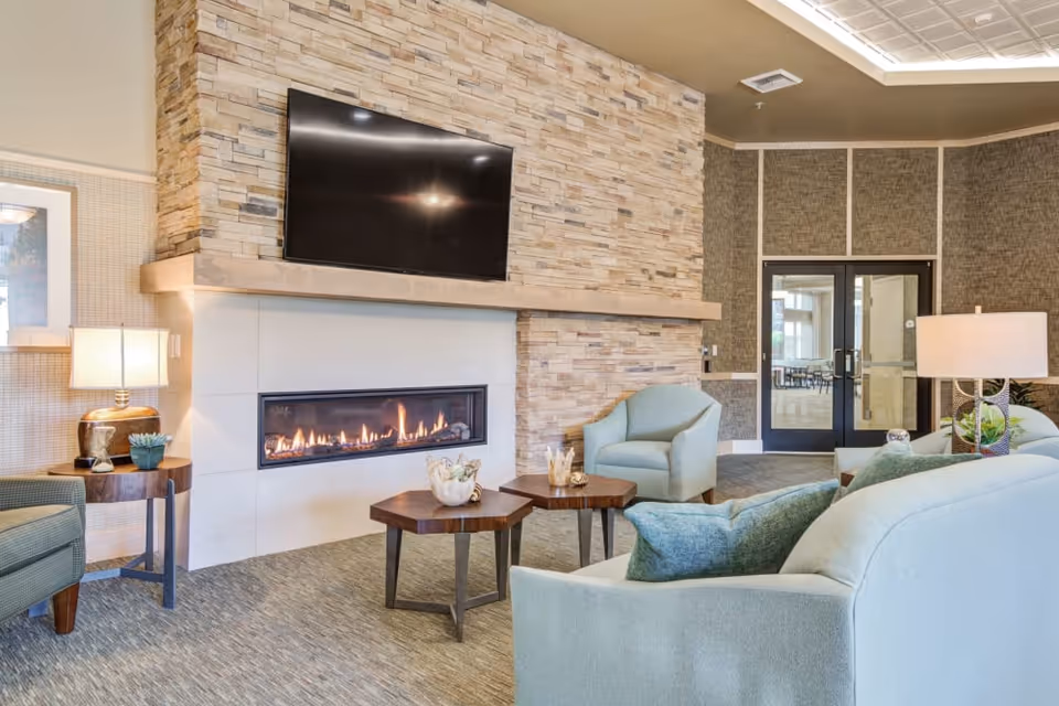 A cozy living room area with a modern stone fireplace and a flat-screen TV mounted above it. The room features light blue upholstered chairs and sofas, two wooden hexagonal coffee tables with decorative items, a side table with a lamp and a small plant, and a glass door leading to another room.