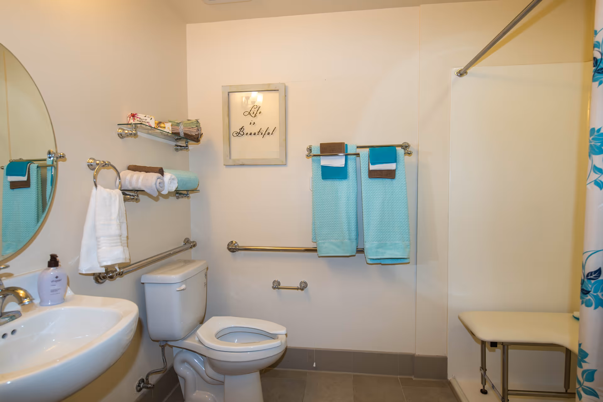 A clean and well-lit bathroom featuring a white toilet, a white sink with a soap dispenser, and multiple towel racks holding turquoise, brown, and white towels. There is a round mirror above the sink, a framed picture on the wall with the text 'Life is Beautiful,' and a shower area with a floral shower curtain and a small bench.