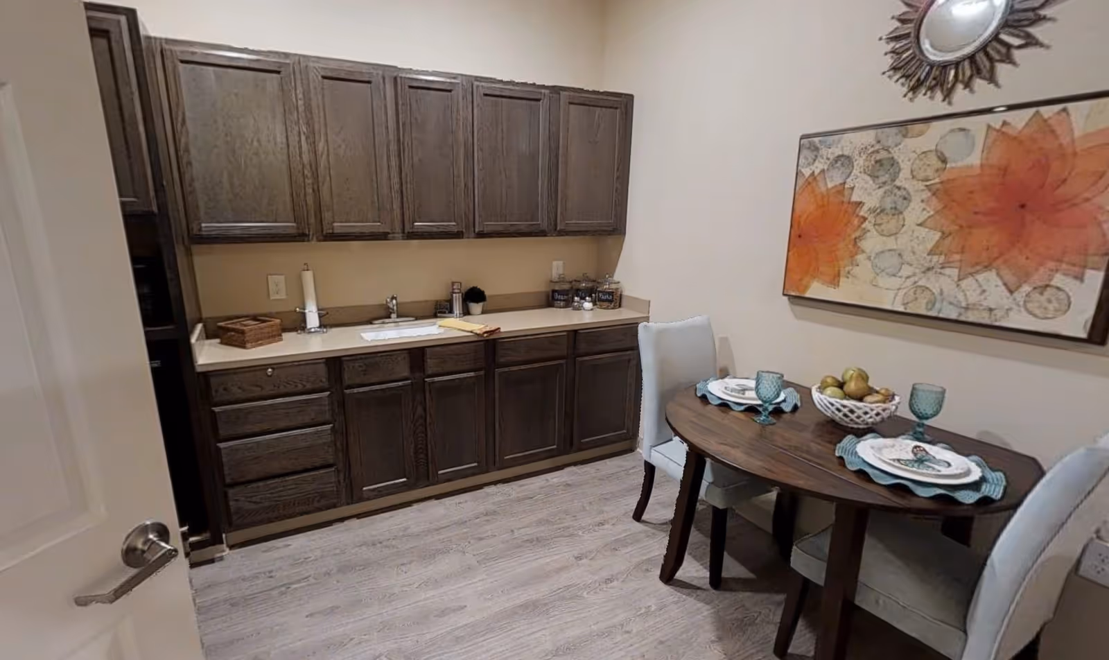 Small dining area with a round wooden table set for two with plates, glasses, and a fruit basket. Adjacent to the table is a countertop with dark wooden cabinets above and below, a sink, and some kitchen items. The walls are light-colored, and there is a decorative wall mirror and a large floral artwork with orange flowers.