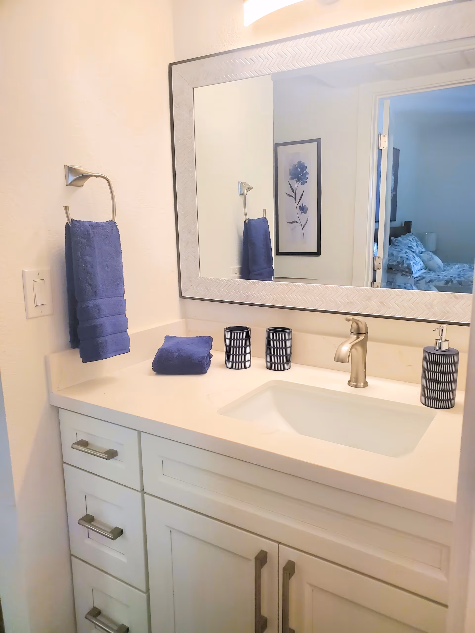 A bathroom vanity with a white countertop and sink, silver faucet, and white cabinetry with silver handles. On the countertop are two blue and white patterned cups and a matching soap dispenser. A folded blue towel is placed on the counter, and a blue hand towel hangs on a silver towel ring mounted on the wall. A large rectangular mirror with a textured white frame is above the sink, reflecting a framed floral artwork and a glimpse of a bedroom in the background.