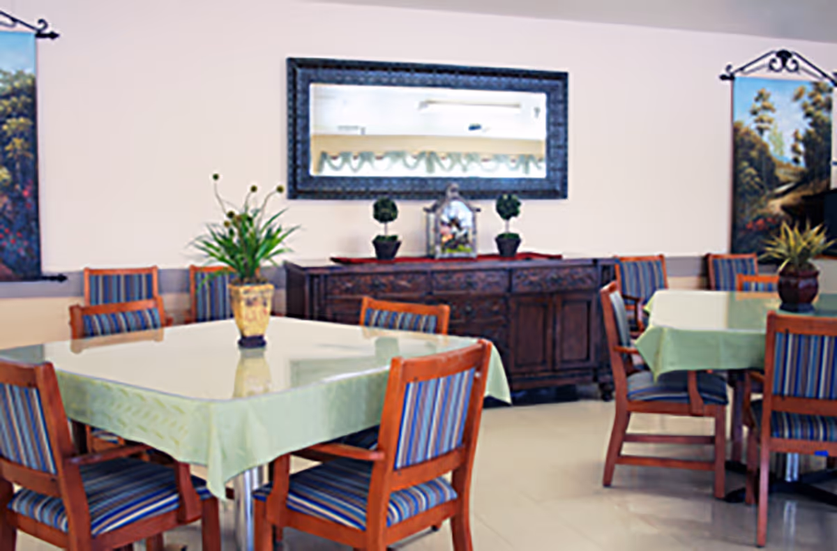 A dining room with tables covered in light green tablecloths, surrounded by wooden chairs with blue striped cushions. There are plants in vases on the tables and a wooden sideboard against the wall with decorative items and a large mirror above it. Two landscape paintings hang on the walls.