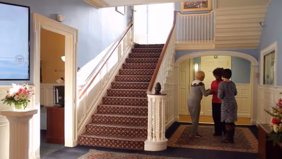 Interior view of a nursing and rehabilitation center showing a carpeted staircase with ornate white railings, three women standing and conversing near a doorway, floral arrangements on pedestals, and light blue walls with white wainscoting.