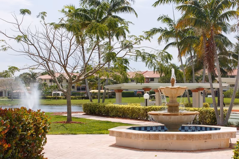 Outdoor view of a landscaped area with a tiered stone water fountain in the foreground, palm trees, a small lake with a water jet, and buildings with red roofs in the background under a partly cloudy sky.