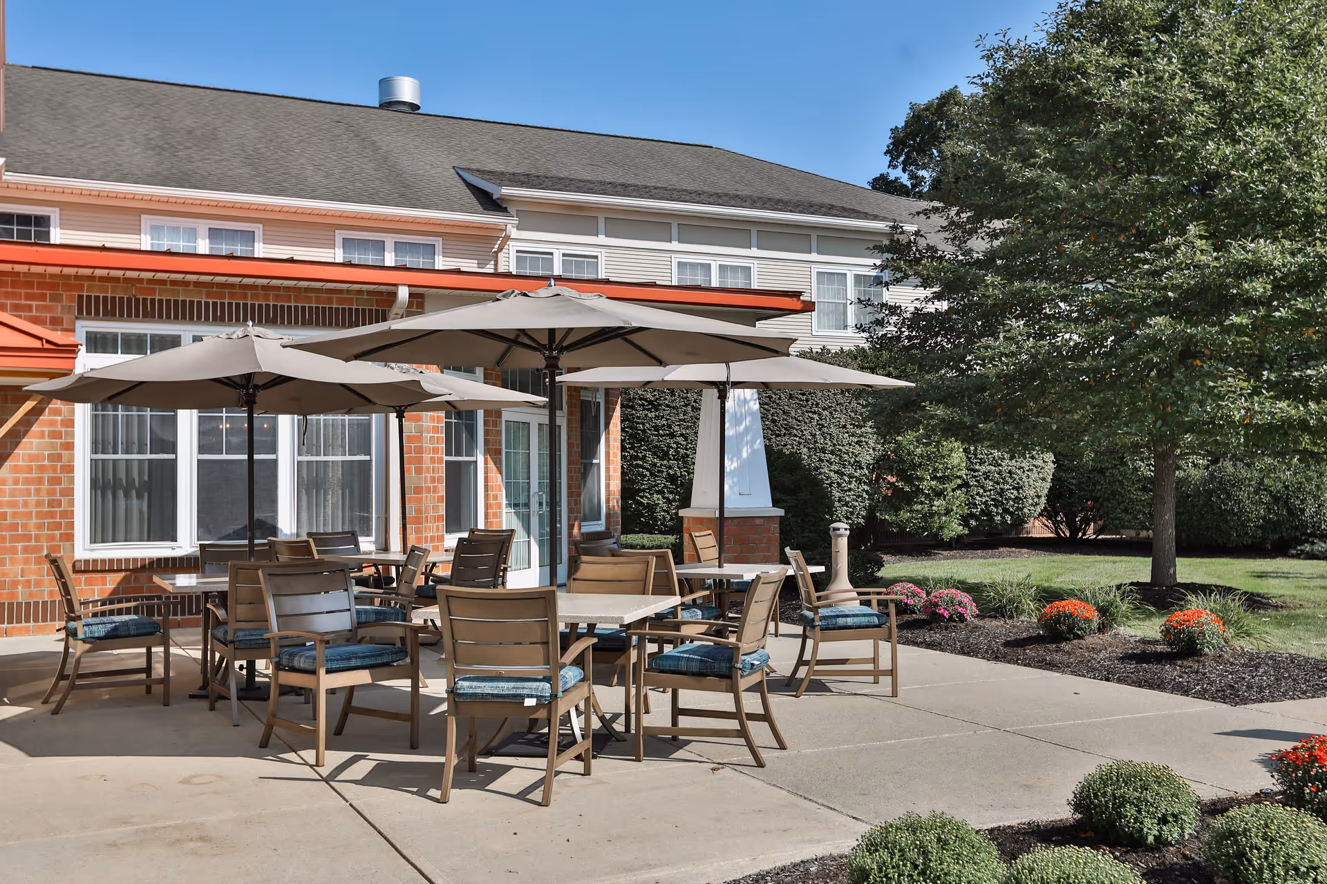 Outdoor patio area at a senior living facility with multiple tables and chairs under large beige umbrellas, surrounded by landscaping with bushes, flowers, and a tree, adjacent to a brick and siding building under a clear blue sky.