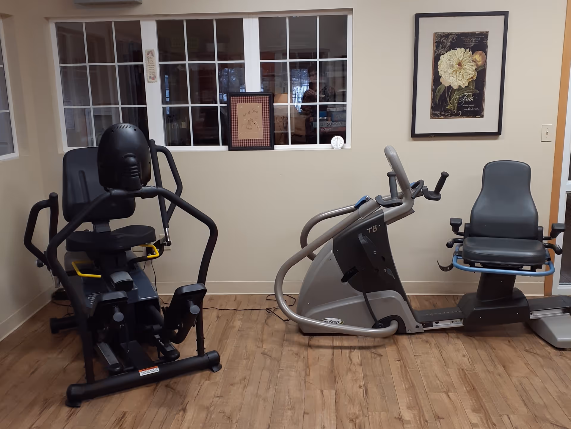 A small exercise room with two pieces of fitness equipment designed for seated workouts. The room has wood flooring, beige walls, a window with multiple panes, and a framed floral artwork on the wall.